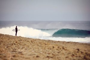 Surfer standing on rocky beach watching a perfect barreling wave at Coxos surf spot in Ericeira, Portugal.