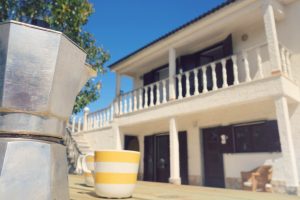 Italian moka pot and striped mug on a wooden table with Moka Surf House guesthouse facade in the background, representing the relaxed atmosphere of the Home Page.