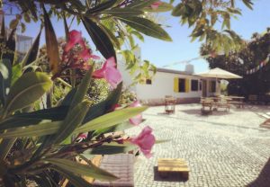 Courtyard at Moka Surf House seen through blooming pink flowers, with outdoor seating and yellow shutters in the background.