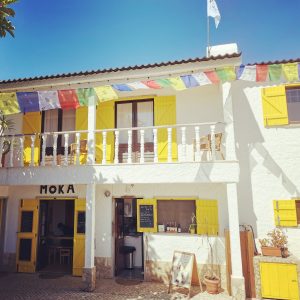 Front view of Moka guesthouse in Ericeira with bright yellow shutters, Tibetan prayer flags, and a small outdoor café area.