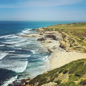 Aerial view of a beach in Ericeira with waves rolling in, surfers in the water, and cliffs lining the coast