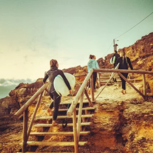 Surfers walking barefoot up wooden stairs on rocky cliffs in Ericeira, carrying their boards after a session.