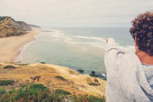 Surf guide pointing at the waves from a cliff above São Julião beach in Ericeira, Portugal, during a surf guiding session.