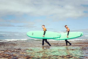 Two surf students in wetsuits walking with soft-top surfboards along the shoreline during a surf school session in Ericeira.