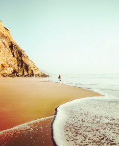 Lone surfer walking along a wide sandy beach with dramatic cliffs and soft waves in Ericeira, Portugal.