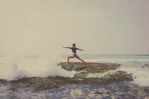 Woman in warrior pose practicing yoga on a rocky shore as ocean waves crash around her during a surf and yoga retreat in Ericeira.